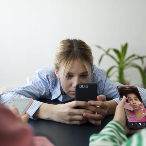 Três meninas estão sentadas à mesa usando seus celulares. A garota ao centro, de blusa listrada azul, parece entediada enquanto segura seu smartphone com as duas mãos. À frente, duas outras meninas manuseiam celulares, e uma delas assiste a um vídeo curto com uma mulher falando. Ao fundo, há uma planta e móveis simples. A imagem transmite uma sensação de excesso de tempo de tela e falta de interação social presencial.